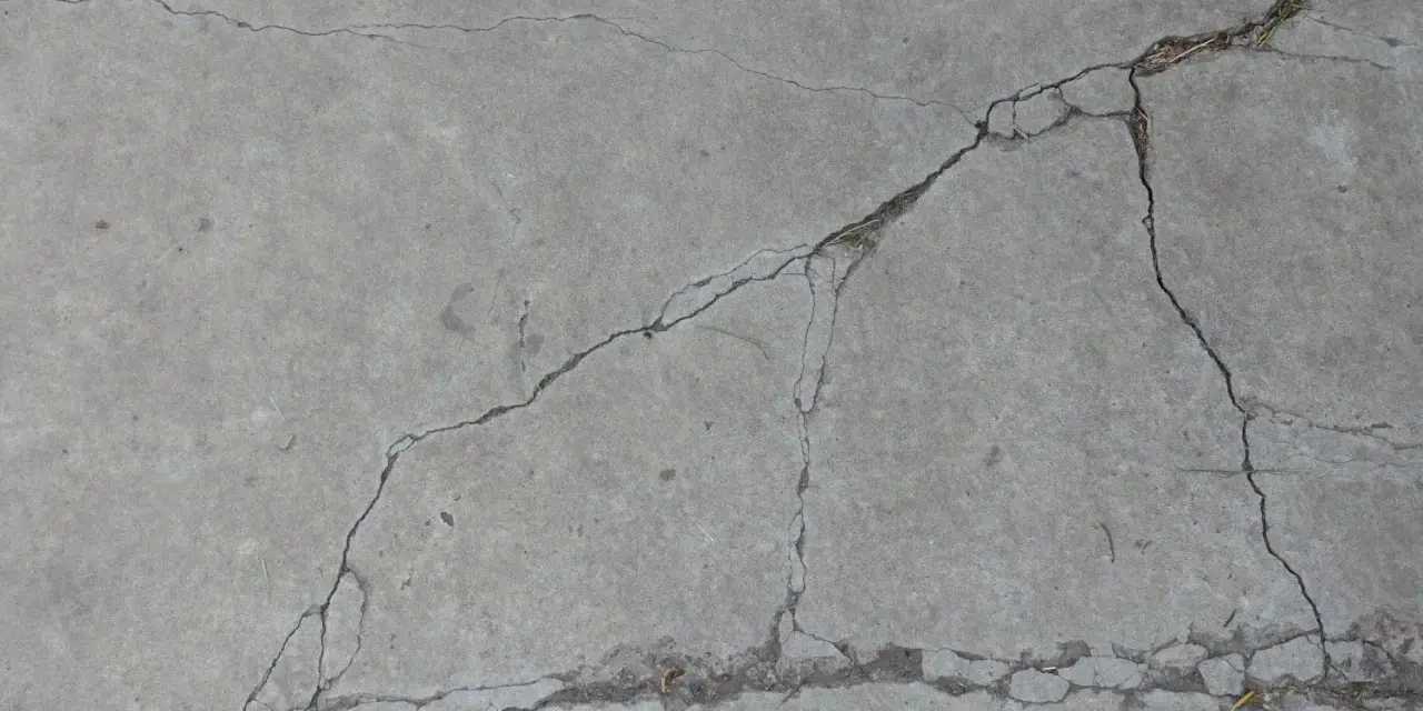 Popękany beton, jakby po długim okresie podlewania, z widocznymi szczelinami i drobnymi źdźbłami trawy.
