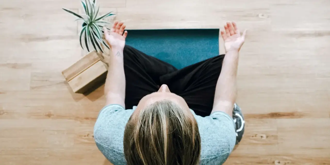 Person mediterar på en yogamatta, en grön växt och träblock bredvid. Perfekt för avkoppling på arbetsplatsen.