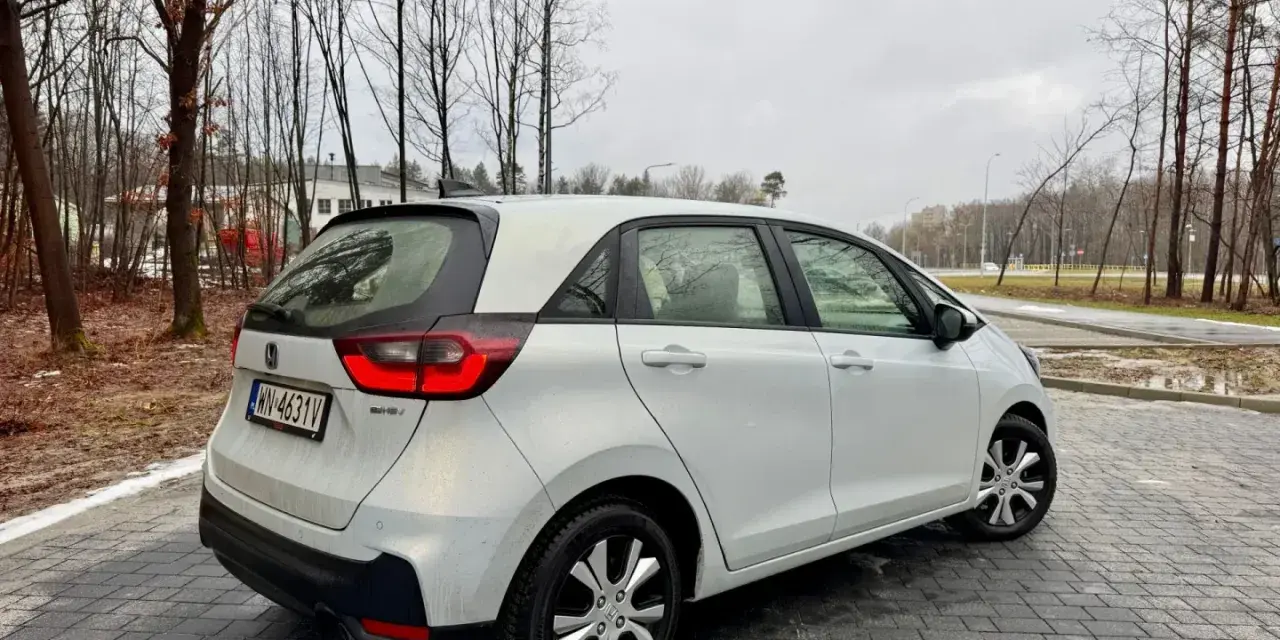 Biały Honda Fit stoi na mokrej, brukowanej nawierzchni. W tle drzewa i budynki.