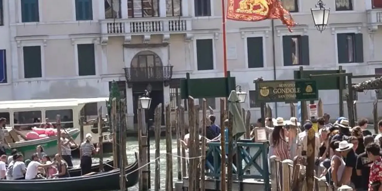 Un gruppo di innamorati a Venezia attende una gondola, con un vaporetto sullo sfondo e il leone di San Marco che sventola.