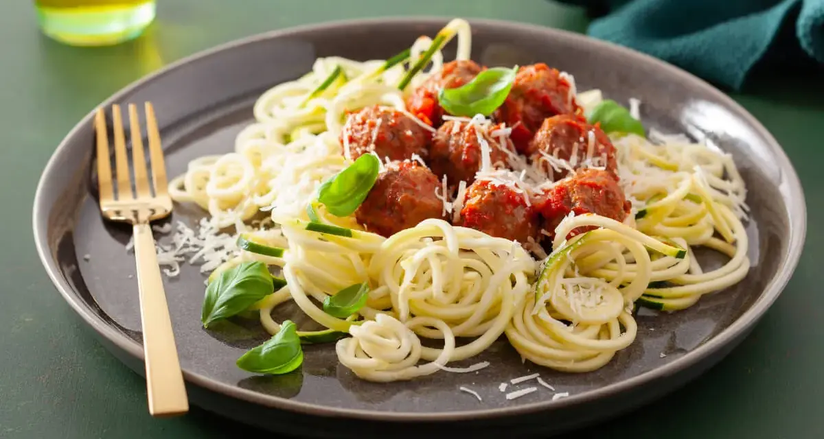Pyszna keto kolacja: klopsiki w sosie pomidorowym na cukinii spaghetti, posypane parmezanem i listkami bazylii. Złoty widelec czeka.