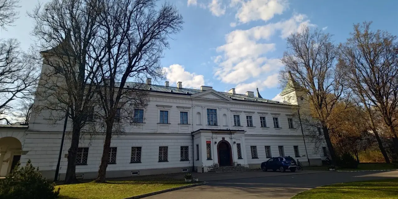 Pałac w Falentach, biała, okazała budowla z wieżyczką, otoczona drzewami o nagich gałęziach, pod błękitnym niebem z chmurami.