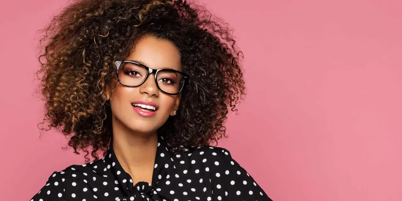 Une jeune femme aux cheveux bouclés porte un maquillage éclatant et des lunettes noires, souriant sur fond rose.