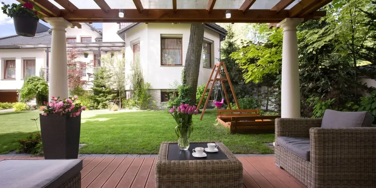 Drewniana pergola z przezroczystym dachem, tworząca zacienione miejsce do wypoczynku na tarasie. W tle ogród z huśtawką i piaskownicą.