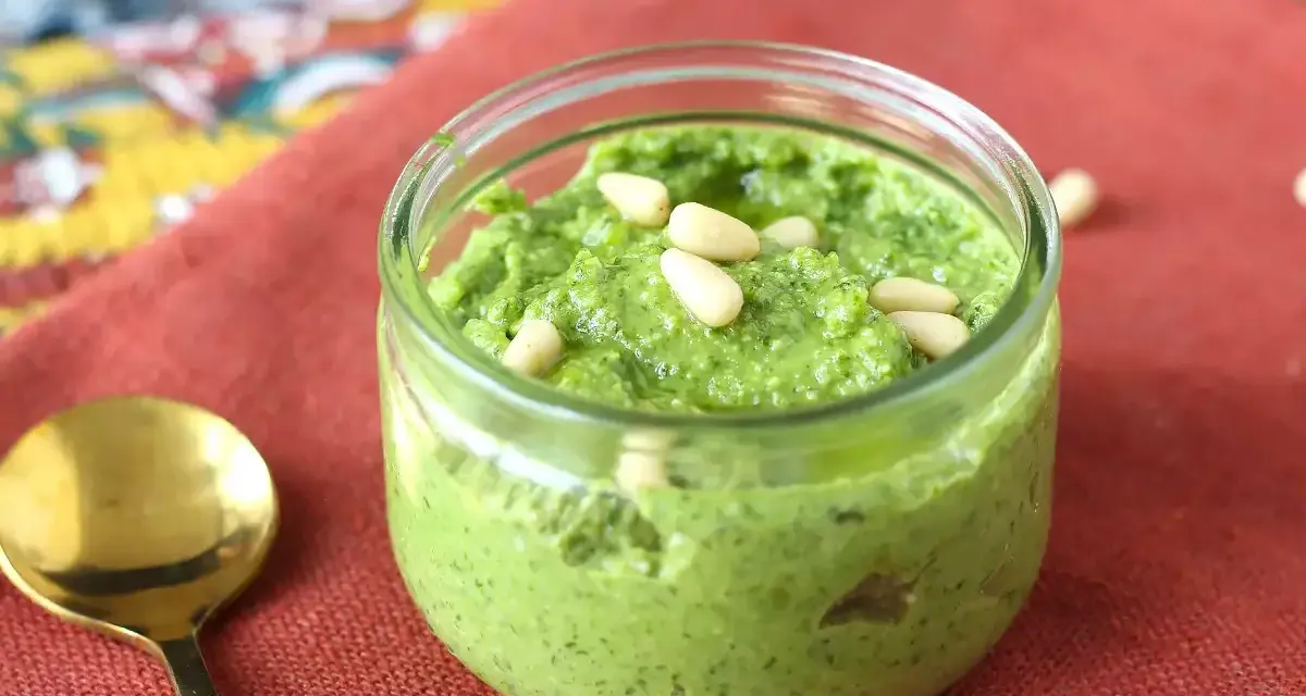 Frisches Rucola Pesto in einem Glas, garniert mit Pinienkernen. Daneben eine goldene Löffel.