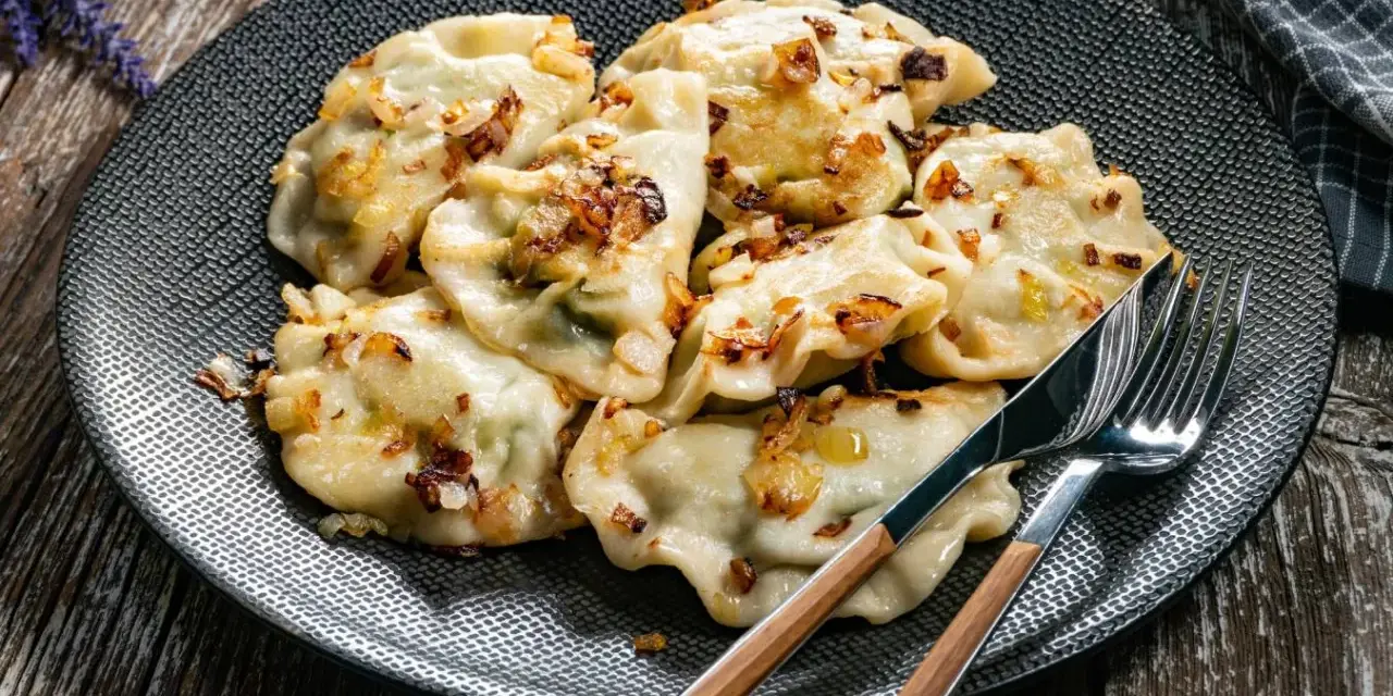 Talerz pełen ugotowanych pierogów z podsmażoną cebulką, obok sztućce.