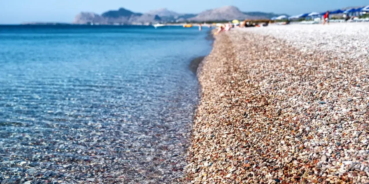 Rodos: turkusowe morze obmywa kamienistą plażę, w tle góry i plażowicze. Idealne miejsce na wakacje.