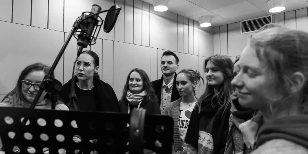 Grupa osób w studiu nagraniowym, przygotowująca podkładanie głosu do bajek. W tle mikrofon studyjny.