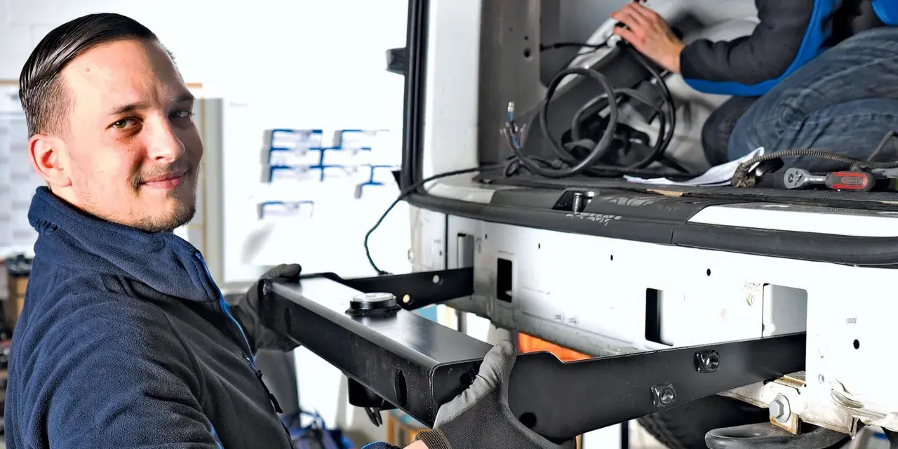 Mechanik z uśmiechem montuje hak w aucie. W tle widać drugą osobę pracującą przy instalacji.