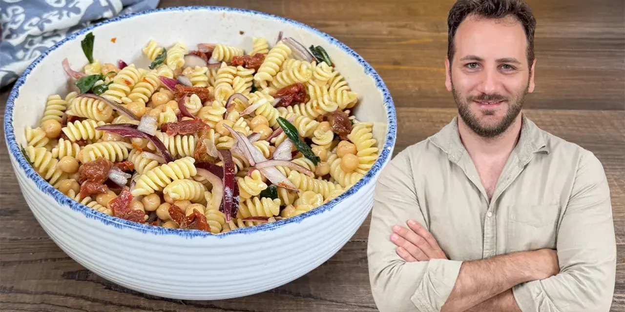 Un uomo sorride accanto a una ciotola di pasta fredda con ceci, pomodori secchi e cipolla rossa.