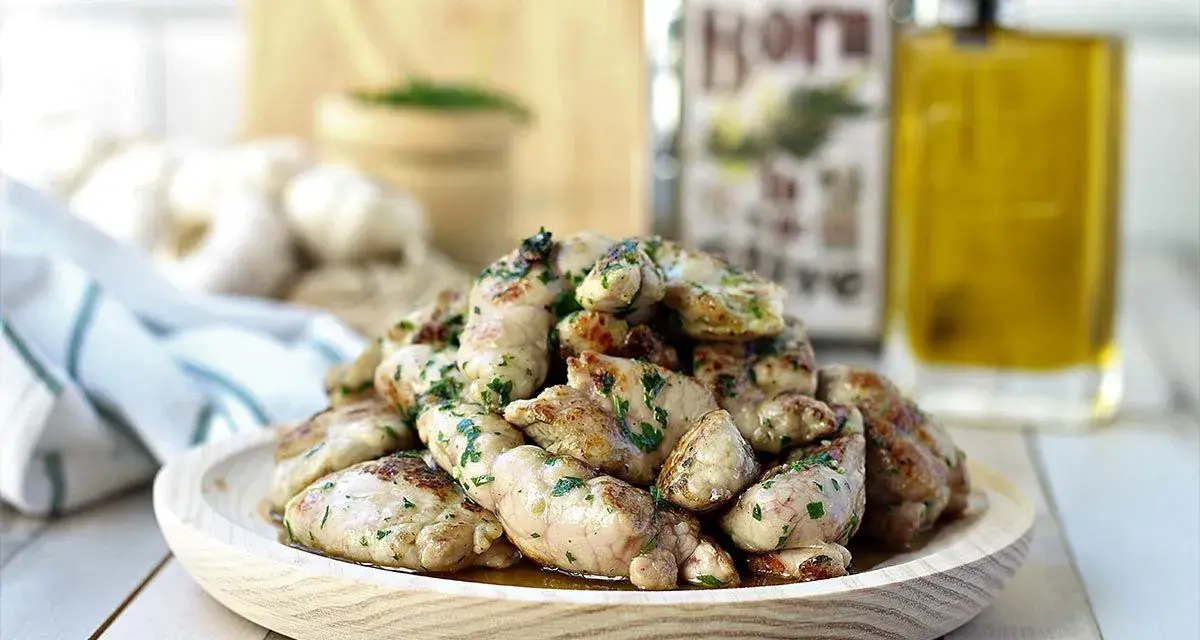 Deliciosas mollejas de cordero salteadas con perejil fresco, servidas en un plato de madera. Al fondo, aceite de oliva y ajos.