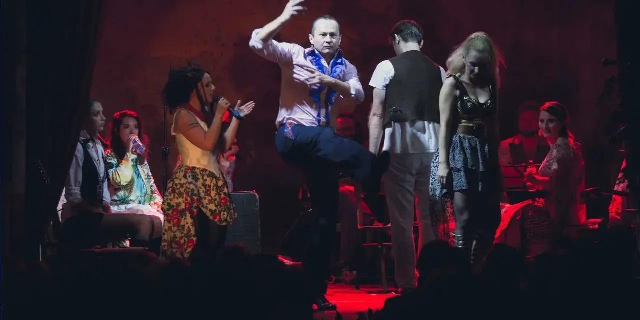 Scena teatralna, gdzie mężczyzna tańczy, a kobiety śpiewają. Atmosfera jak po pożarze w burdelu, pełna emocji i dramatyzmu.
