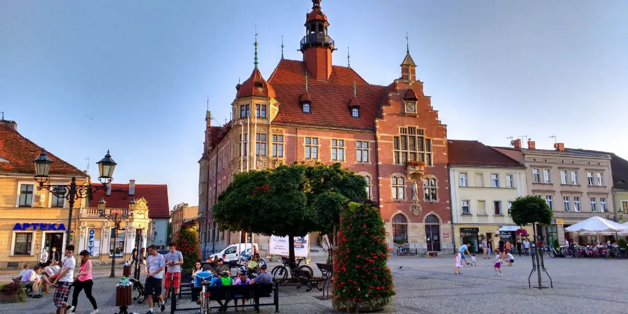 Rynek w Tarnowskich Górach tętni życiem. Ludzie spacerują, siedzą na ławkach, a dzieci bawią się na bruku. W tle widać zabytkowy ratusz.