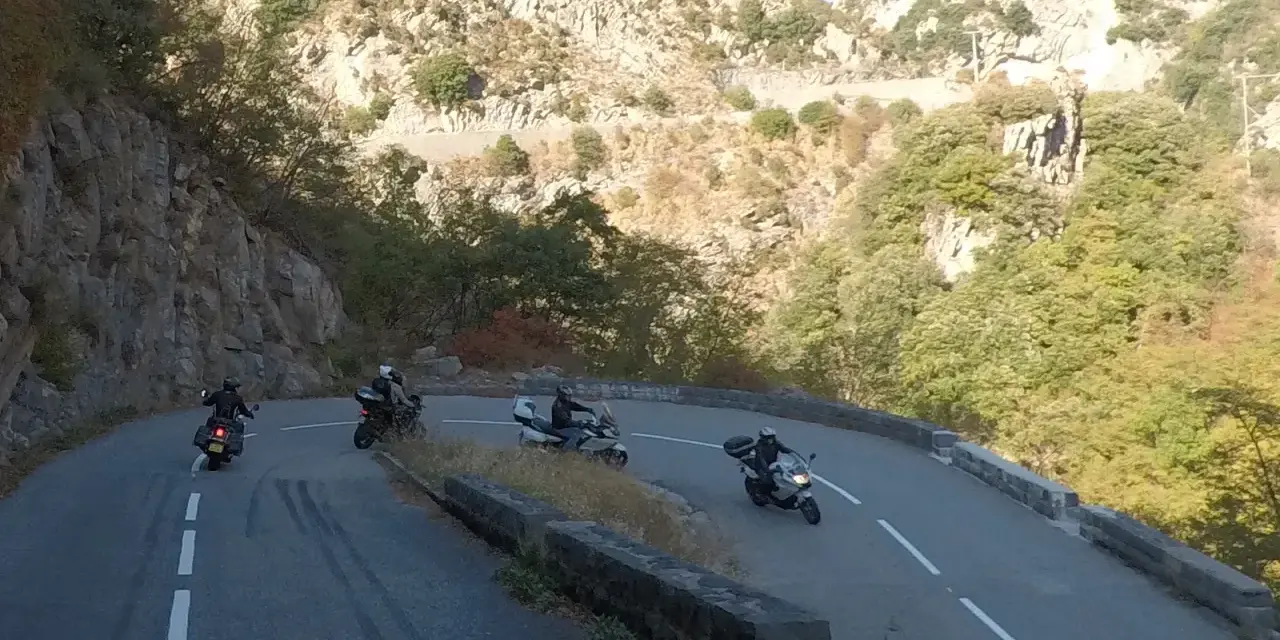 Motociclisti affrontano le curve del Col de Turini, immersi in un paesaggio montano roccioso e verdeggiante.