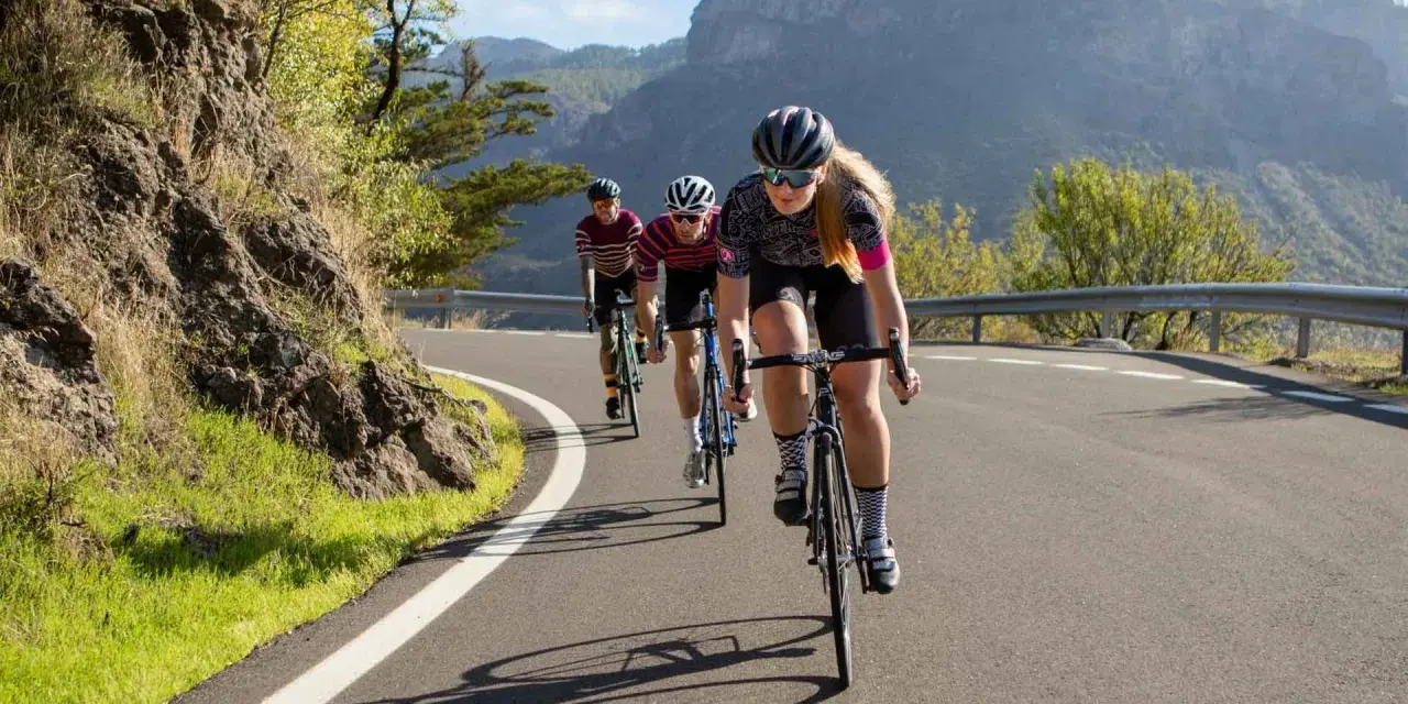 Three cyclists race up a mountain road. The lead cyclist, a woman, is what is a sportive person, focused and determined.