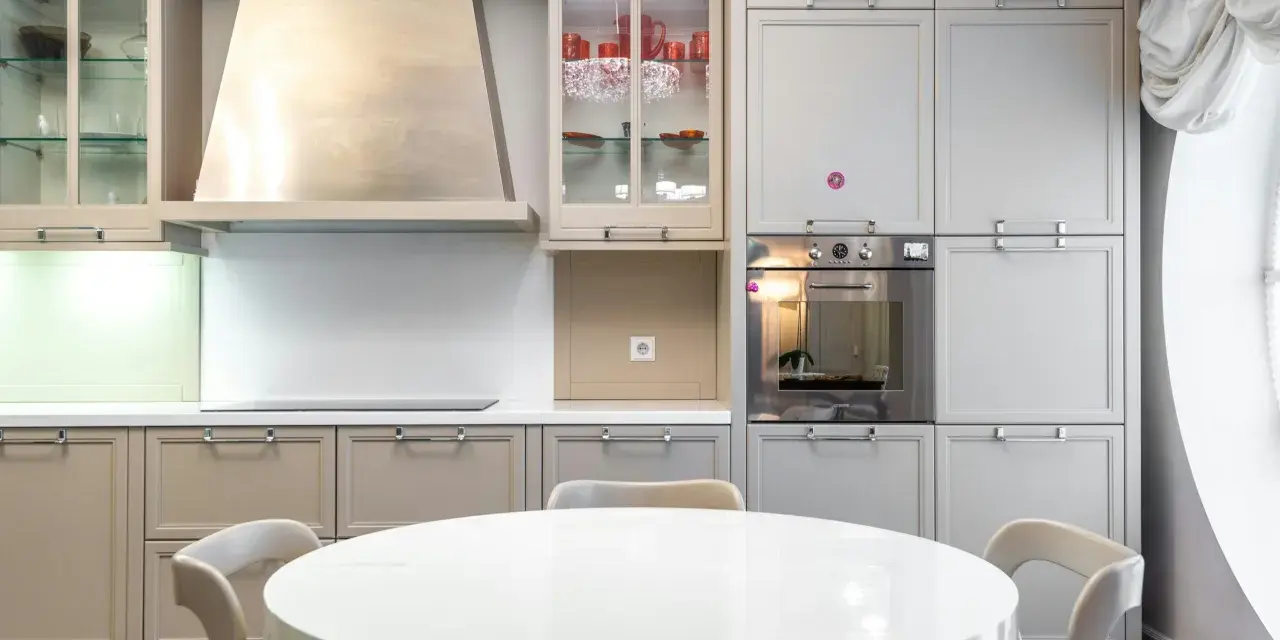 Cocina moderna con mesa redonda blanca y sillas. Gabinetes grises y beige, campana extractora de acero inoxidable. Ideal para ver catálogos de cocinas y precios.