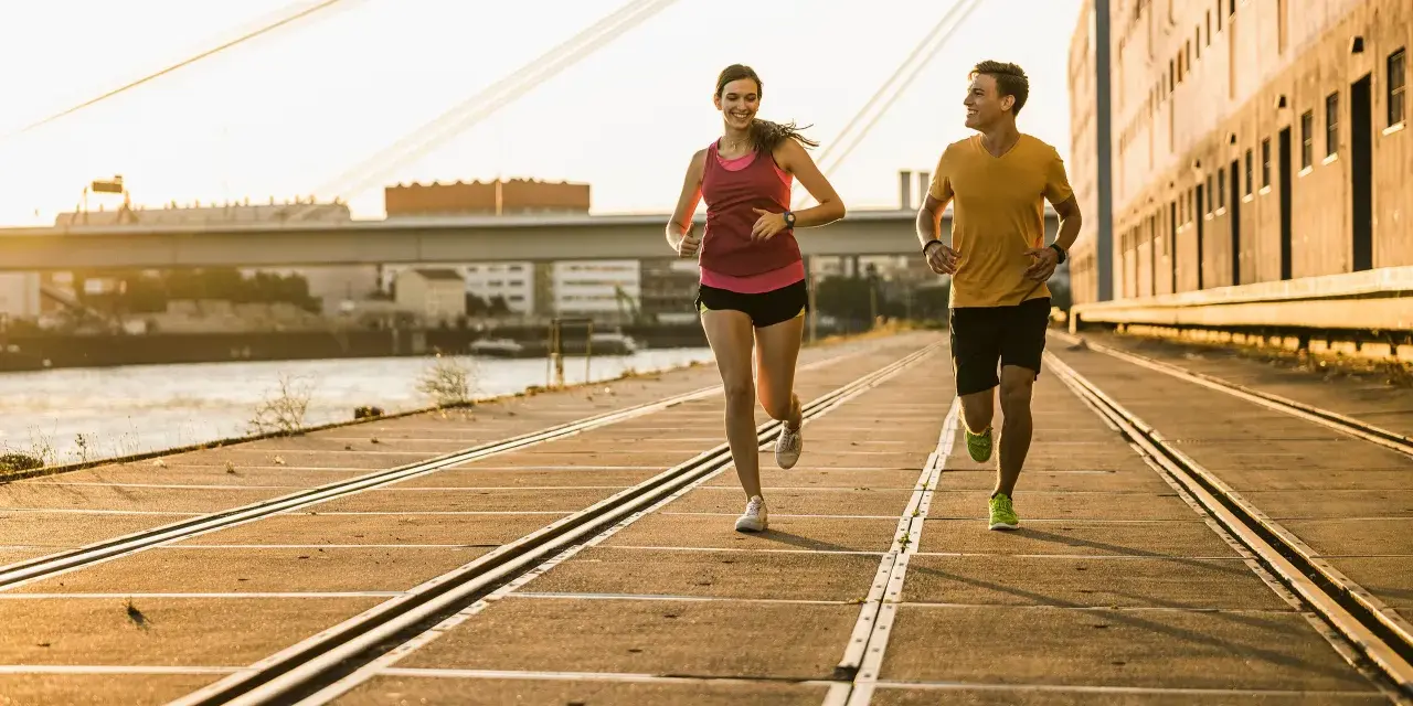 Pareja corriendo al atardecer, demostrando su **endurance crossfit**. El sol dorado ilumina su camino junto al río.