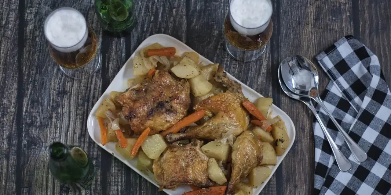 Pollo asado con patatas y zanahorias en una bandeja blanca, con cerveza y servilleta a cuadros.