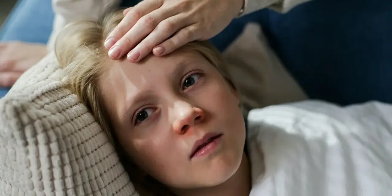 Dziecko z gorączką leży na poduszce, a ręka mamy sprawdza mu czoło. Czy dziecko z gorączką może wychodzić na dwór?