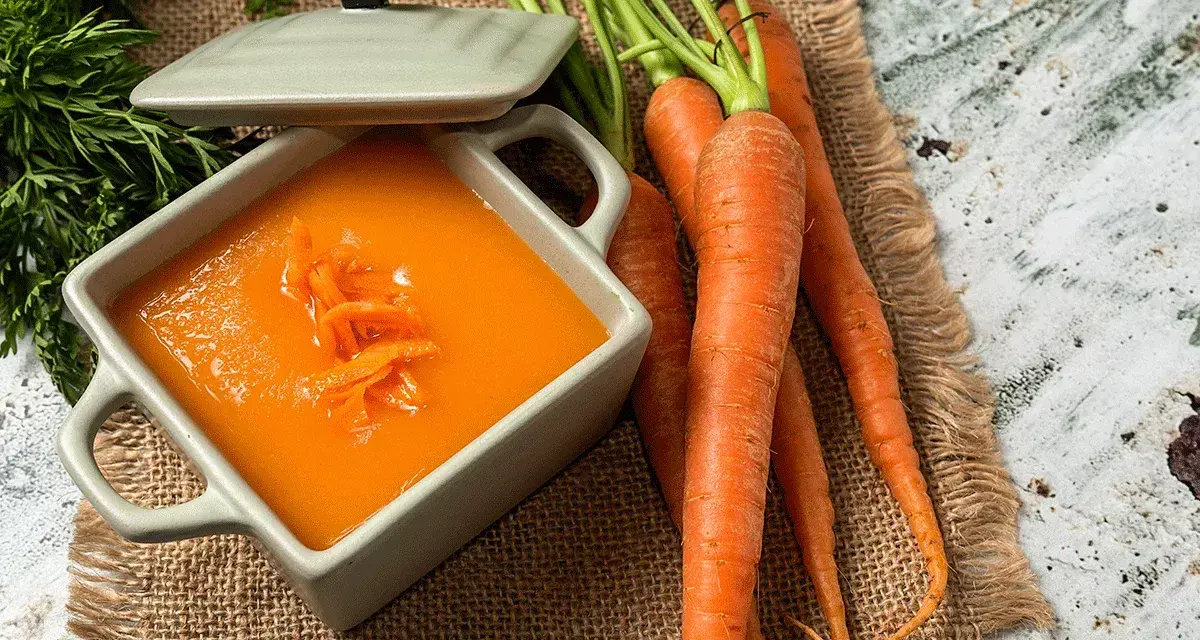 Un tazón de cremoso puré de zanahoria, adornado con hebras de zanahoria rallada, junto a zanahorias frescas y verdes.