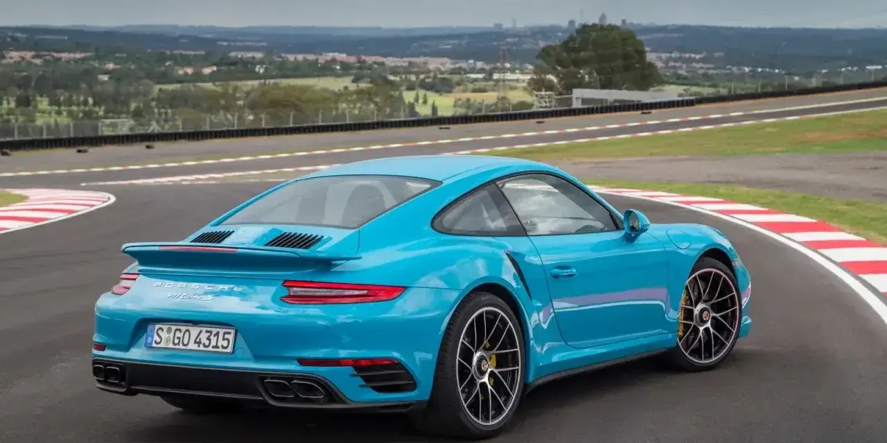 Błękitne Porsche 911 Turbo S na torze wyścigowym, z widokiem na miasto w tle.