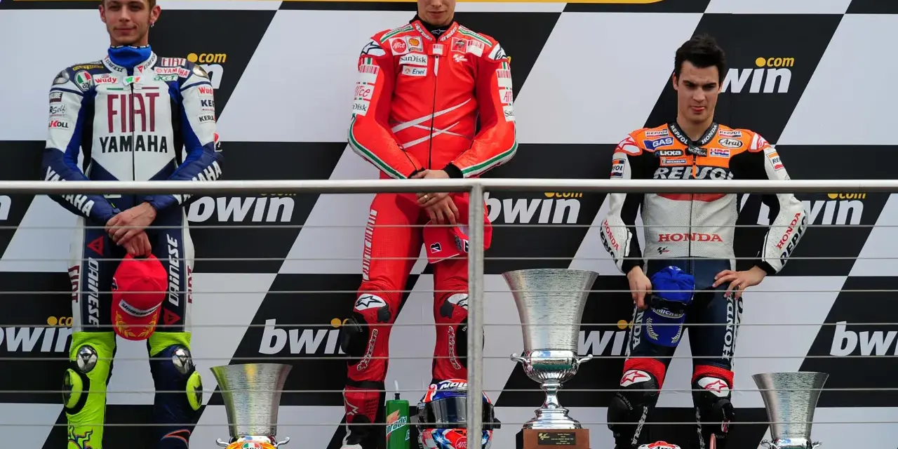Podio con tre motociclisti famosi in tuta: Rossi, Stoner e Lorenzo. Vittoria celebrata con trofei.