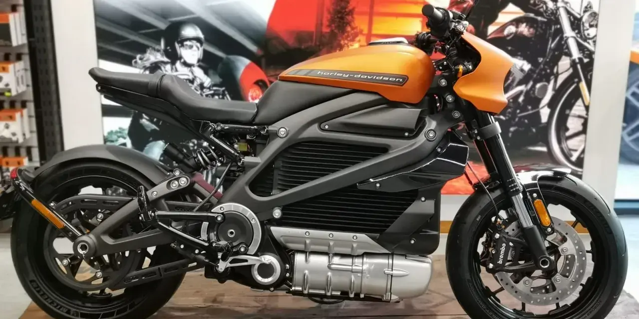 Pomarańczowy harley elektryczny z czarnymi akcentami, prezentowany na drewnianej podłodze, z plakatem Harley-Davidson w tle.