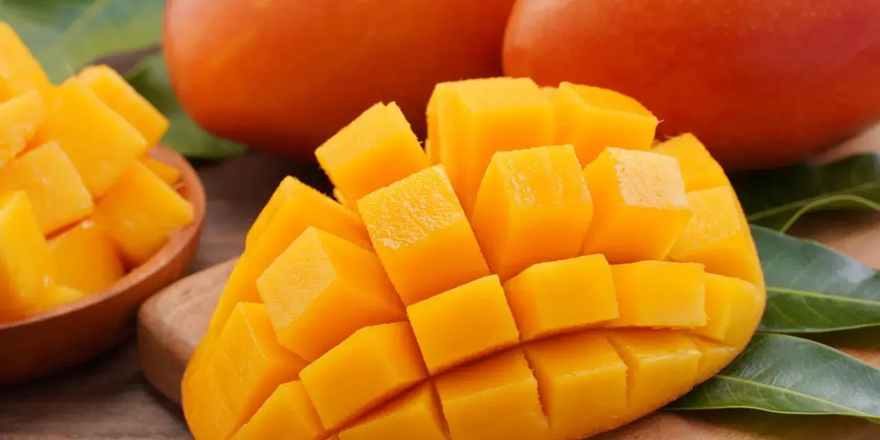 Dojrzałe mango, pokrojone w kostkę, prezentuje swoje soczyste, pomarańczowe wnętrze. Obok leżą całe owoce i liście.