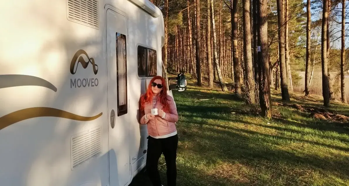 Kobieta z kubkiem kawy opiera się o kampera na leśnej polanie. Wspaniała przygoda w **Finlandii kamperem**!