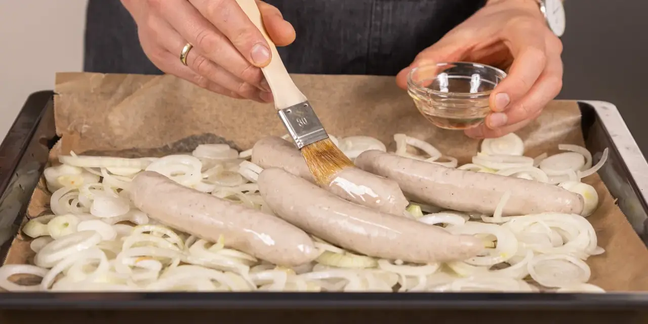Bratwurst im Backofen: Zwiebelringe auf Backpapier, darauf vier rohe Bratwürste. Mit einem Pinsel wird Öl aufgetragen.