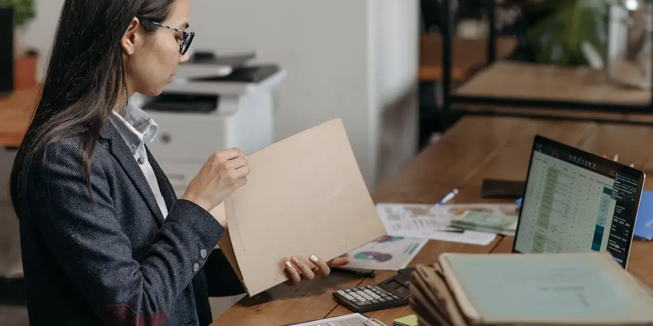 Kobieta w okularach przegląda dokumenty, szukając tytułu prawnego do lokalu. Na biurku leżą też laptop, kalkulator i sterty papierów.