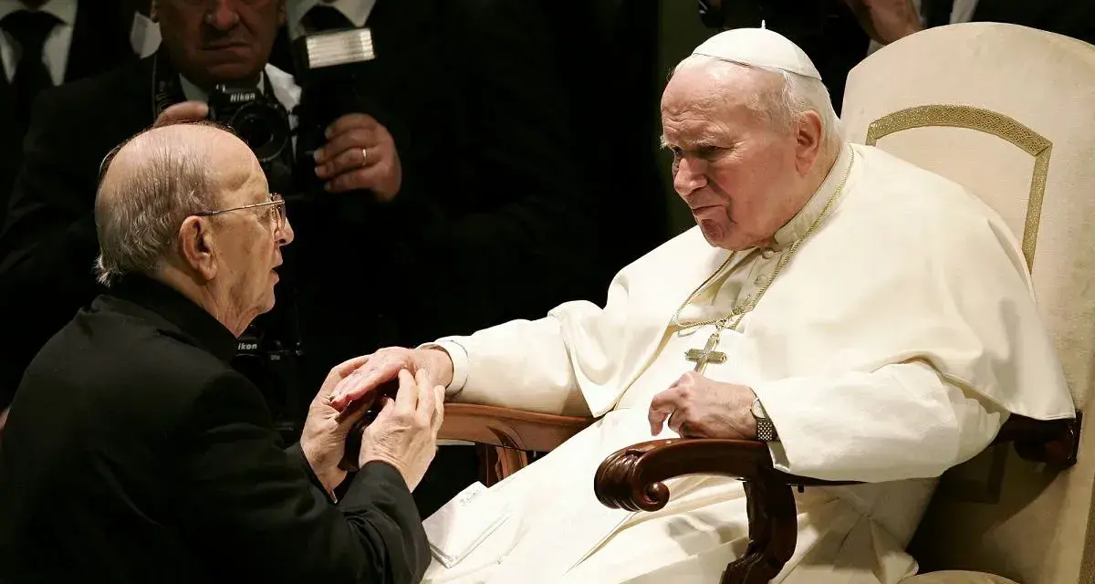 Papież Jan Paweł II w białej szacie, klęczący przed nim mężczyzna w czerni. W tle widać innych duchownych i dziennikarzy, być może z zakonu **jezuitów**.