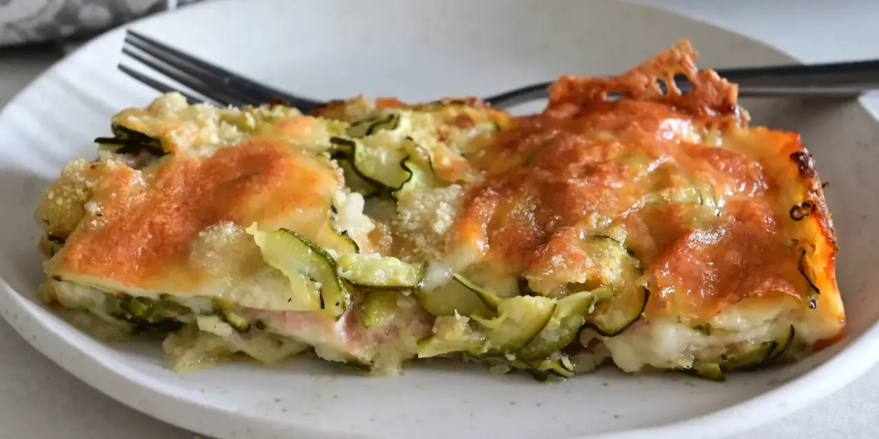 Una fetta di sformato di zucchine dorato e filante, con formaggio fuso e zucchine a rondelle, servito su un piatto bianco.