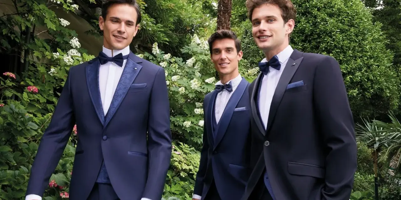 Tre eleganti sposo e testimoni in abiti scuri, sorridono in un giardino lussureggiante.
