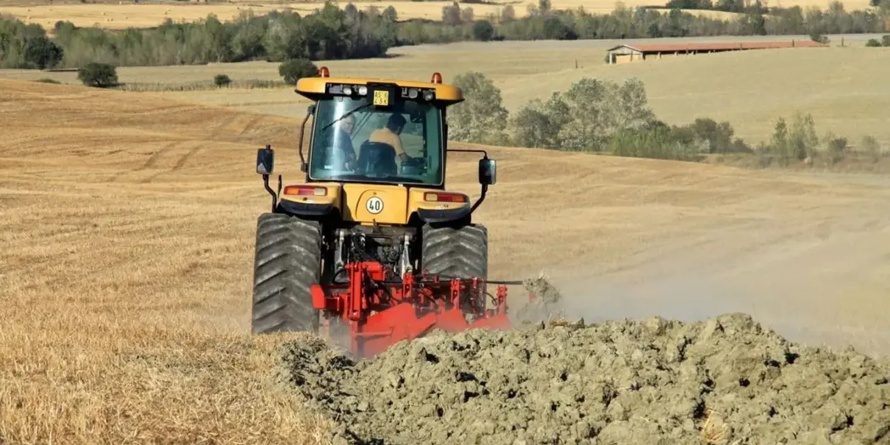 Trattore giallo in azione per l'aratura del terreno, sollevando terra e polvere in un paesaggio rurale collinare.