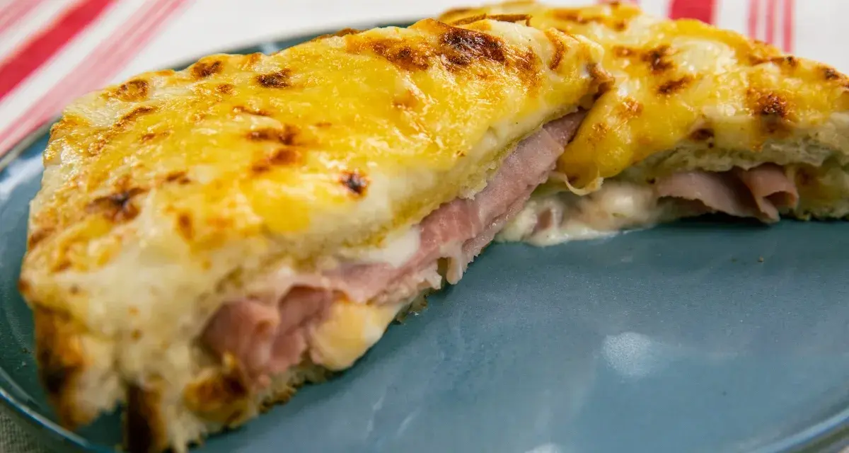 Croque Monsieur gratinado con queso derretido y jamón, servido en plato azul.