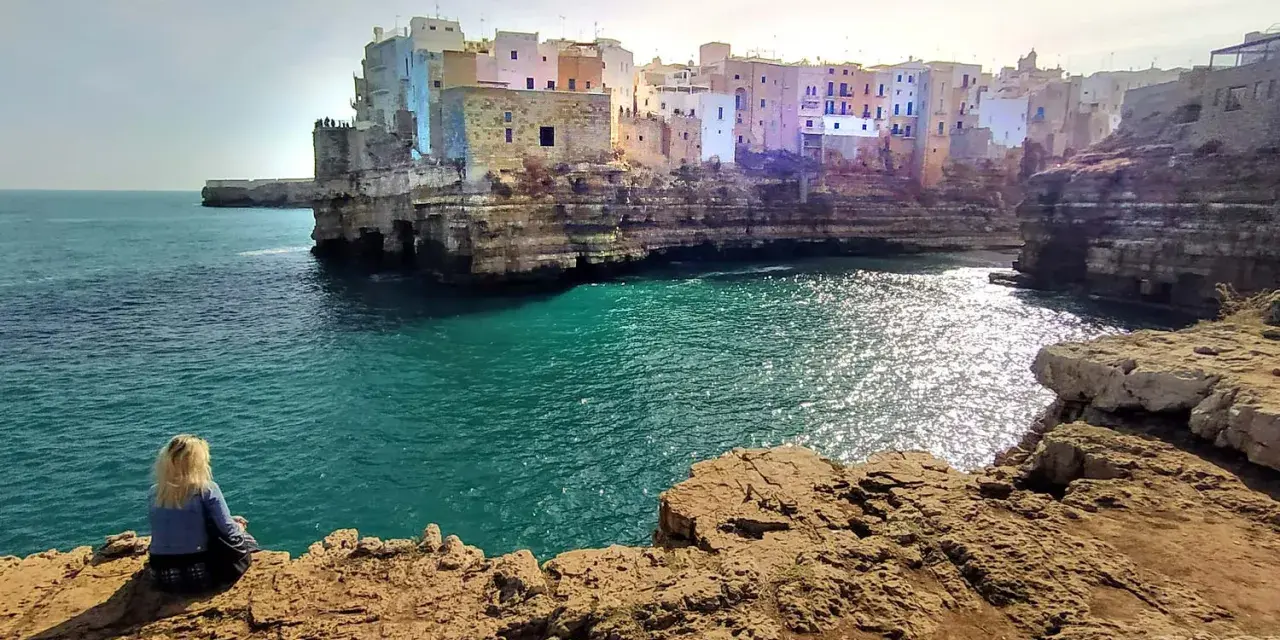 Kobieta siedzi na skale, podziwiając widok na Polignano a Mare. Białe domy wznoszą się na klifie nad turkusowym morzem.