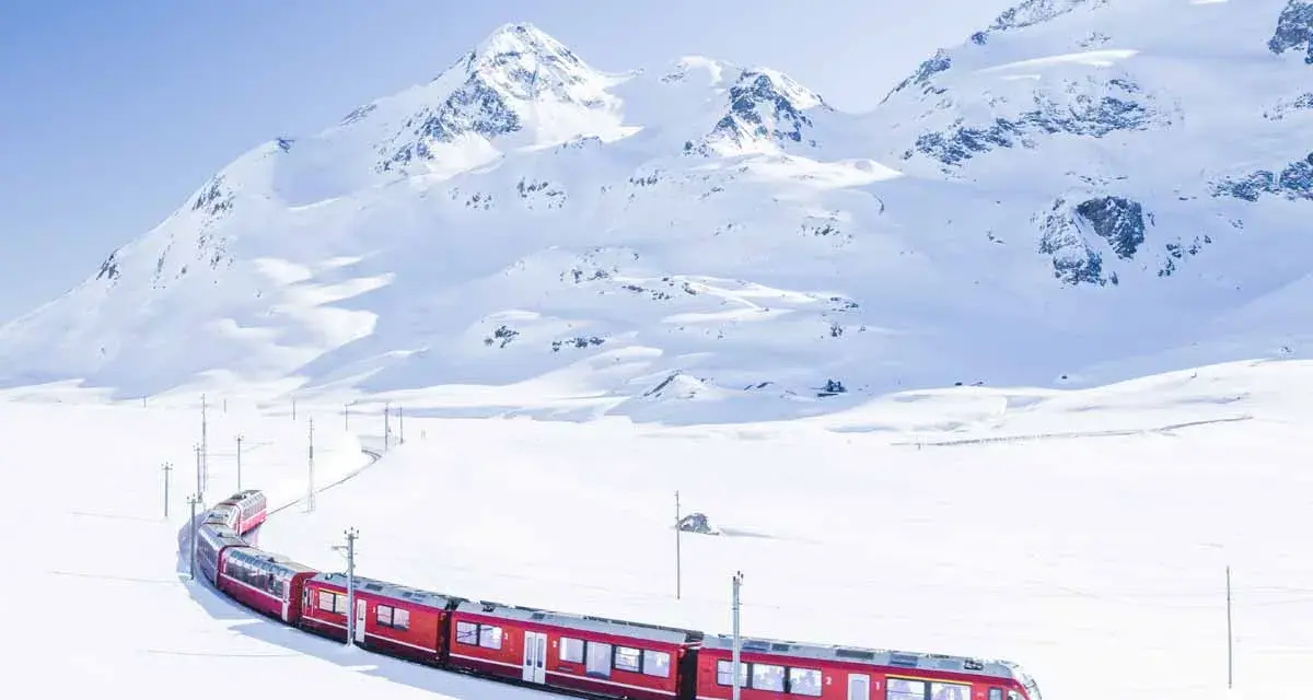 A bright red St. Moritz train winds through a pristine, snow-covered mountain landscape under a clear blue sky.