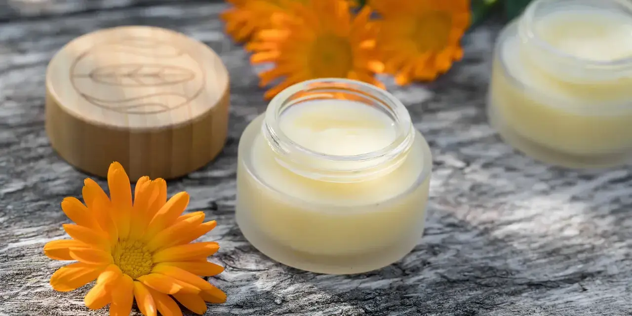 Ringelblumensalbe gegen Falten: Natürliche Pflege für glatte Haut. Kleine Gläser mit Salbe und leuchtend orangefarbene Ringelblumenblüten auf Holz.