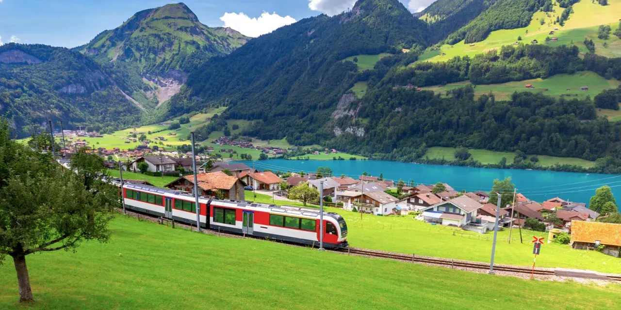 A red and white train travels through a picturesque Swiss village nestled beside a turquoise lake, with lush green mountains in the background. This is the scenic route from Lucerne to Lugano.