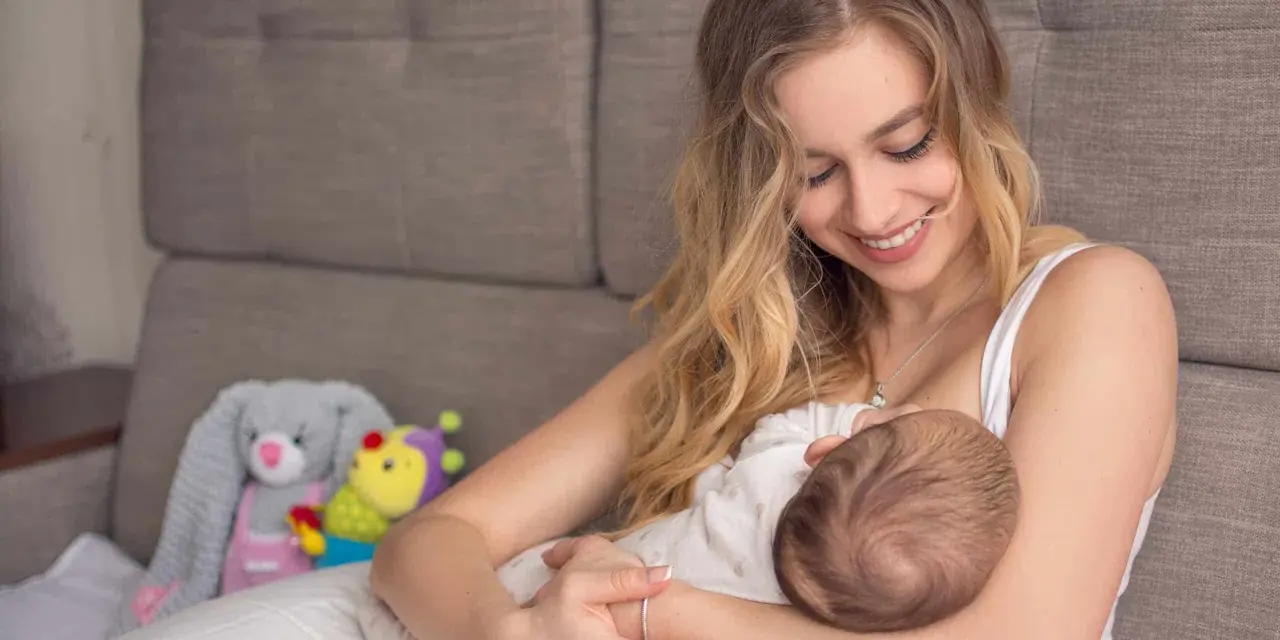 Uśmiechnięta mama karmi piersią dziecko. W tle pluszowe zabawki. Nakładki na pierś do karmienia mogą pomóc w tym intymnym momencie.