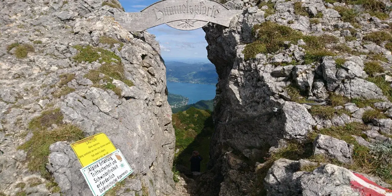 Himmelspforte am Schafberg: Ein Felsentor mit Holzinschrift, das den Blick auf einen See freigibt. Ein Wanderer steht im Durchgang.
