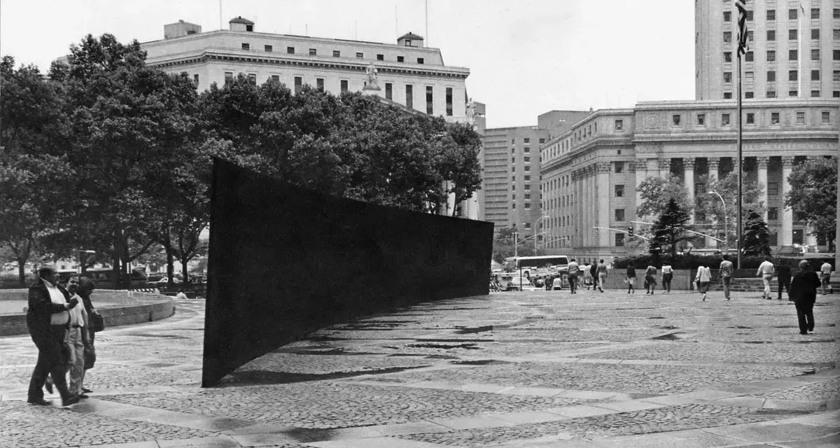 Monumentalne dzieło Richarda Serry, masywna, czarna ściana, dominuje nad placem. Ludzie spacerują wokół, a w tle widać budynki.