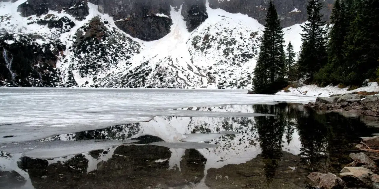 Morskie Oko zimą, zamarznięte, z odbiciem ośnieżonych gór i ciemnych drzew w przejrzystej wodzie.
