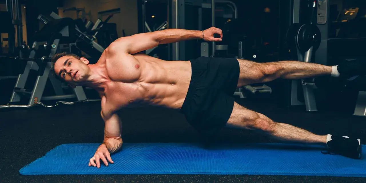 Un homme musclé réalise un gainage planche sur un tapis bleu dans une salle de sport.