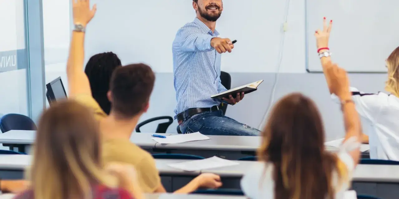 Uśmiechnięty wykładowca z książką w ręku wskazuje na studentów, którzy podnoszą ręce. Czy przed lecz stawiamy przecinek?