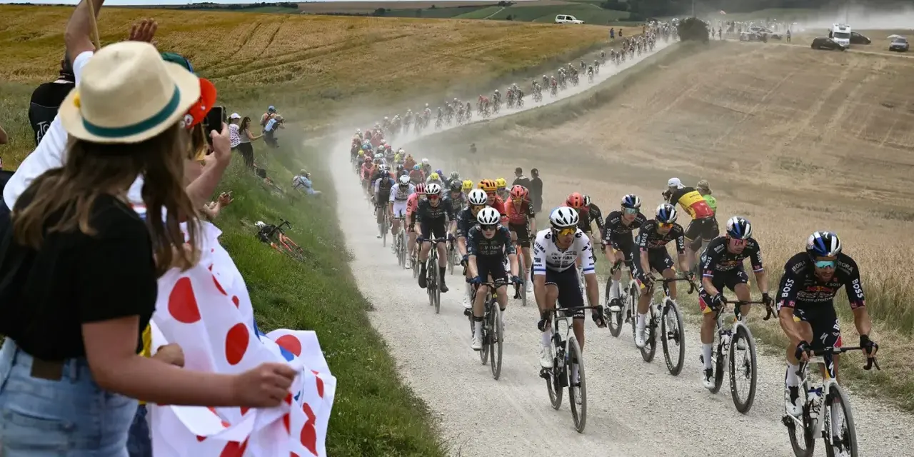 Spectators cheer as a peloton races on a dusty road during the Tour de France watch. Cyclists in colorful jerseys kick up dust in a dramatic scene.