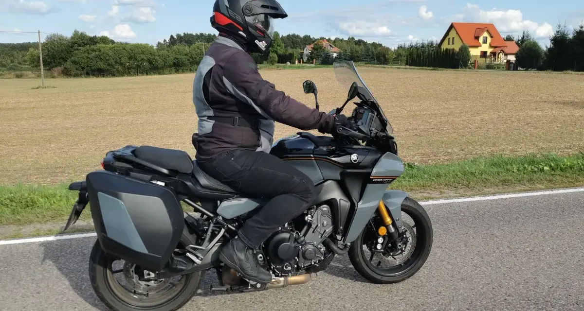 Motocyklista na szarym motocyklu Yamaha Tracer 900, z kuframi, w kasku, jedzie drogą. W tle pola i domy. Yamaha Tracer 900 dane techniczne.