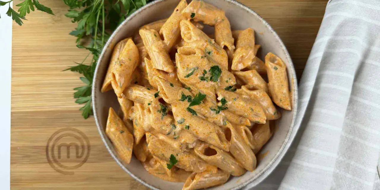 Penne alla vodka cremose e invitanti, guarnite con prezzemolo fresco, servite in una ciotola su un tagliere di legno con il logo MasterChef.