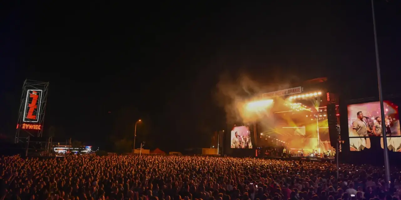 Tłum ludzi na koncercie Żywiec Męskie Granie. Na scenie gra zespół, a na telebimach widać artystów.